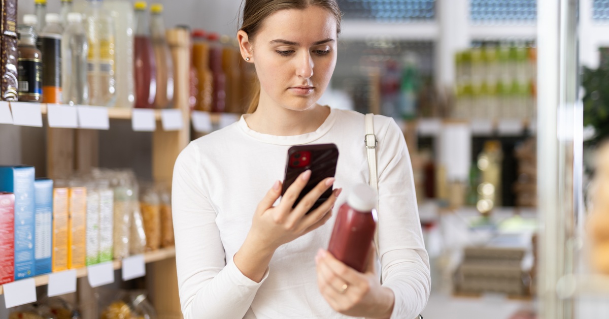 A young female customer stands in the aisle as she takes a photo of the back of a product. She's wearing a white shirt.