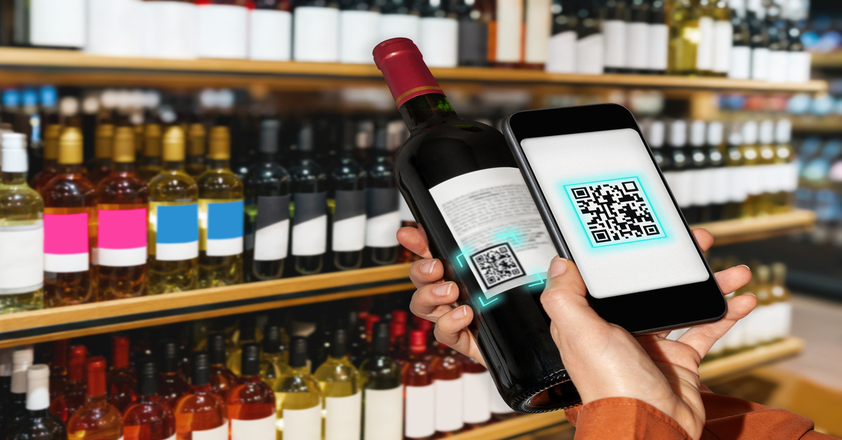A close-up of a person scanning a wine bottle QR code with their phone. There are rows of wine bottles behind them.
