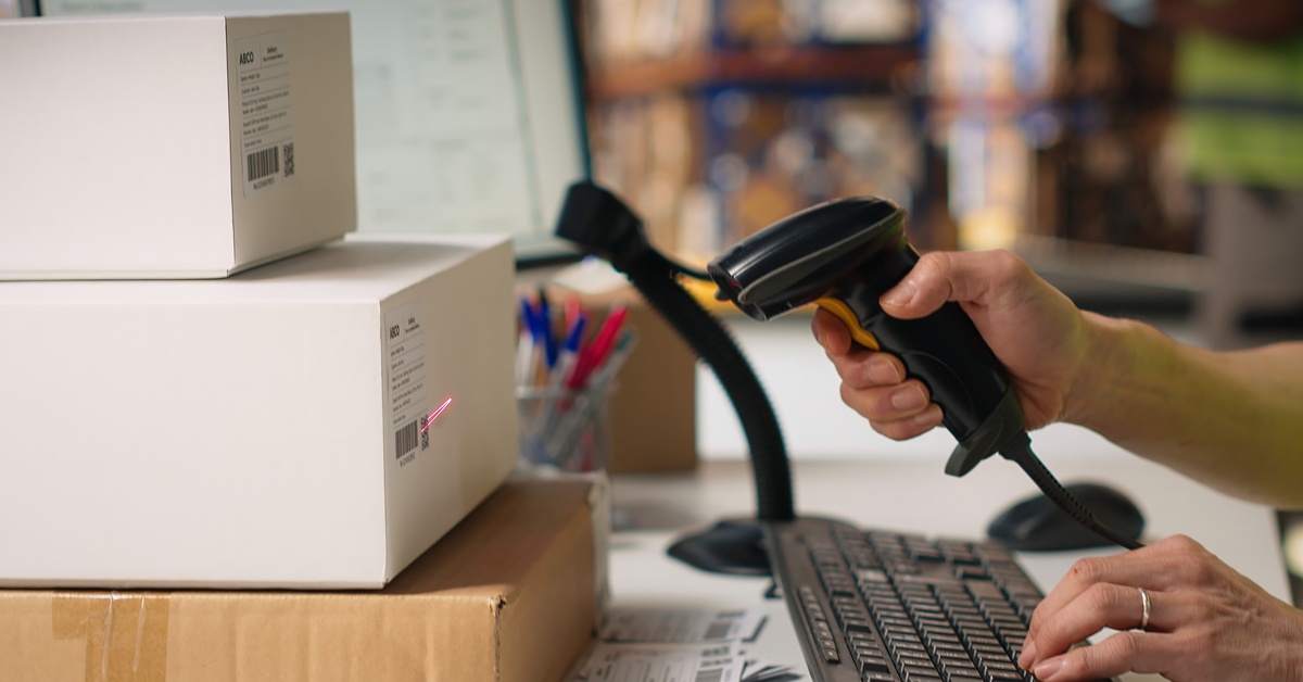 A close-up of an employee's hands at their desk scanning labels on a stack of boxes. The boxes are brown and white.