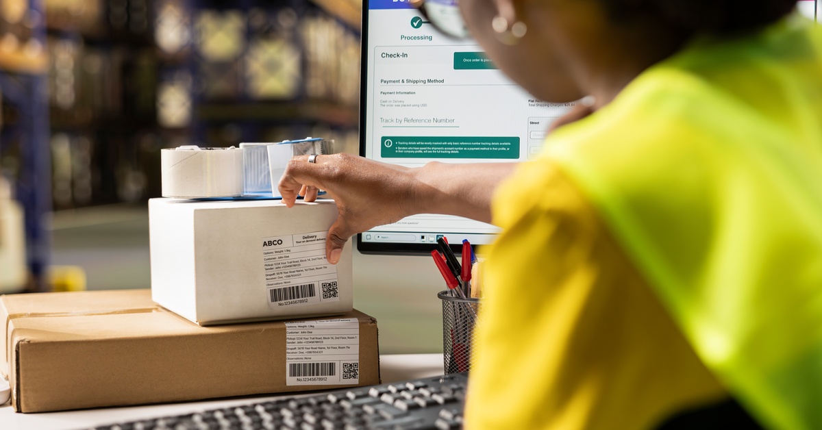 A warehouse employee sits at the workstation in front of the computer monitor, placing a label on a product.