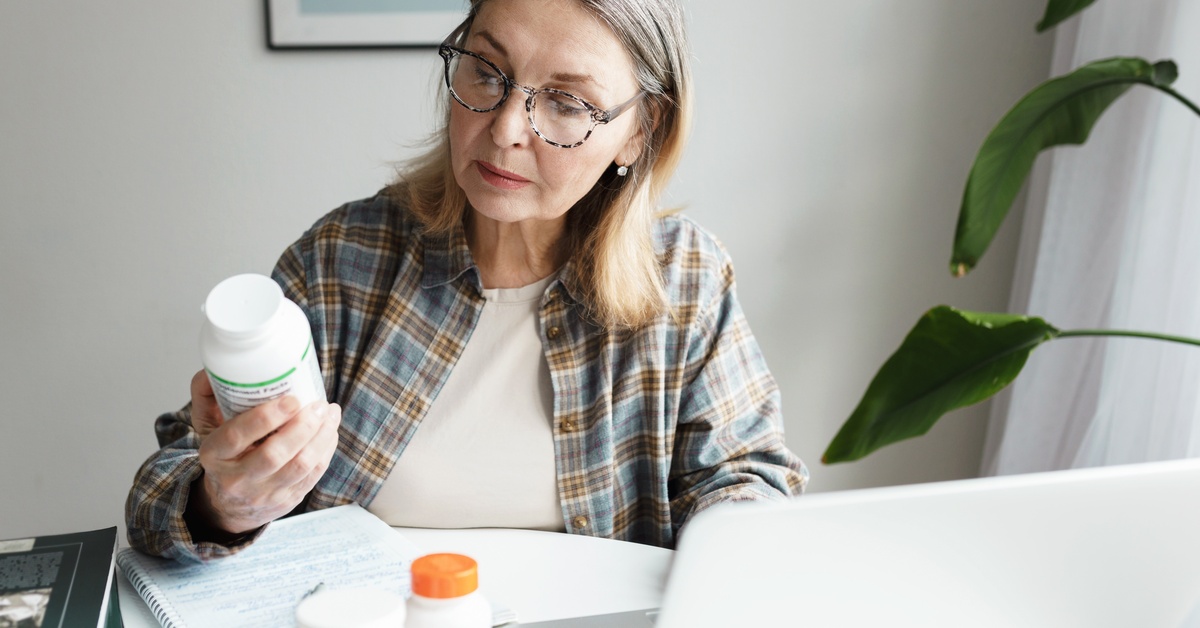 An aging woman wears glasses as she sits at the table with a laptop. She is reading product labels on vitamin bottles.