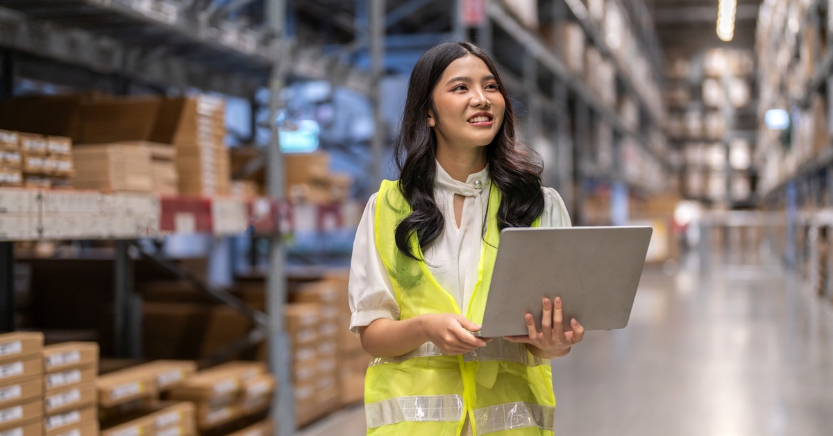A young female warehouse employee wears a yellow safety vest as she holds a laptop to check inventory levels.