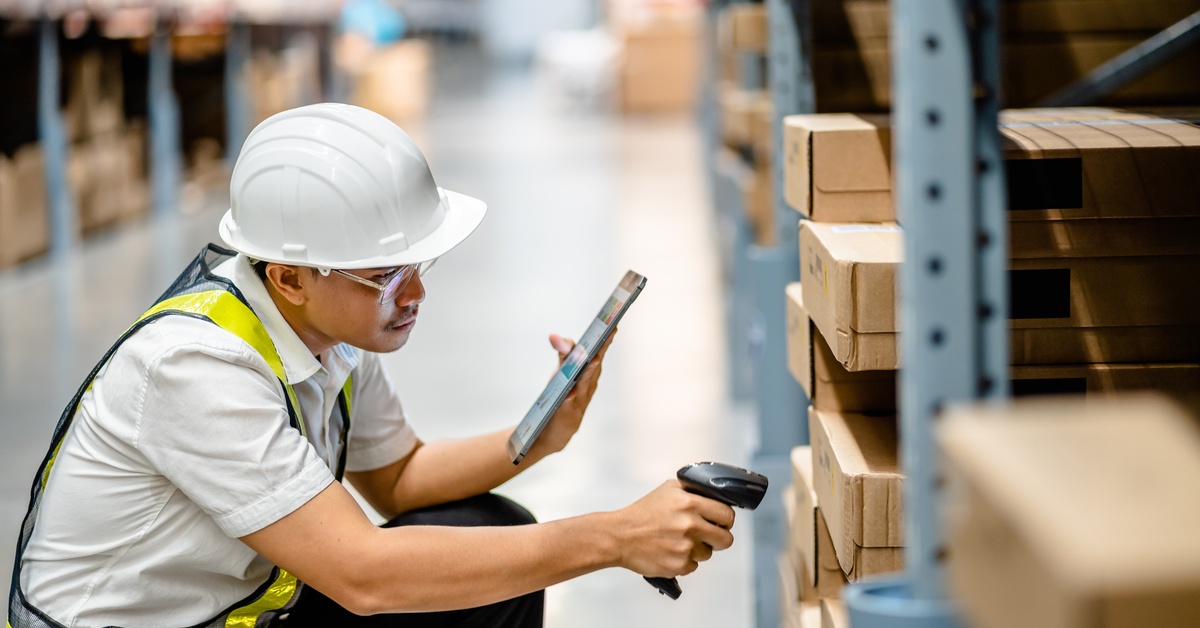 A warehouse employee in a safety vest and hardhat scans packages in the warehouse with a tablet and handheld scanner.