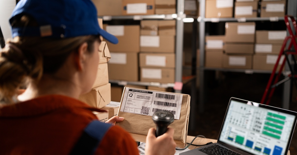 A warehouse employee in a baseball cap and ponytail sits at the desk facing the software, scanning a box label.