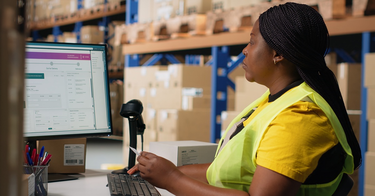 A Black woman employee sits at the desk in a warehouse with the label software system open on the desktop monitor.